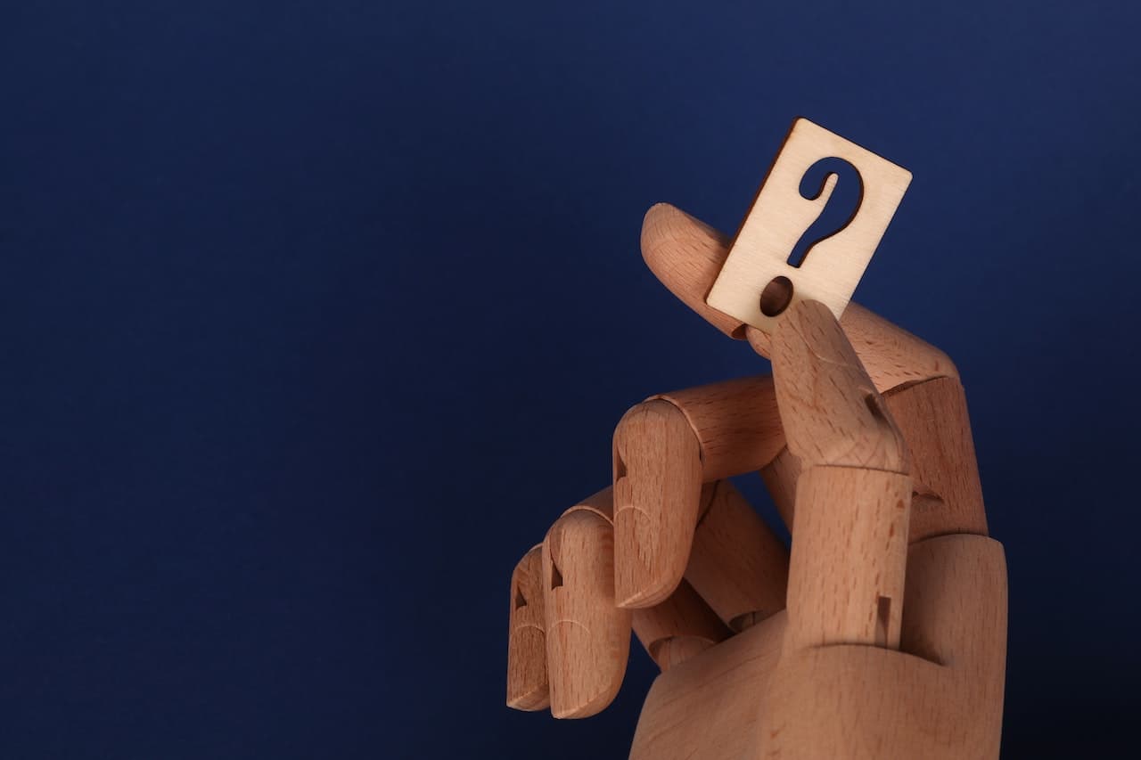 Wooden artist's mannequin hand holding a small wooden tile printed with a question mark, against a dark blue background